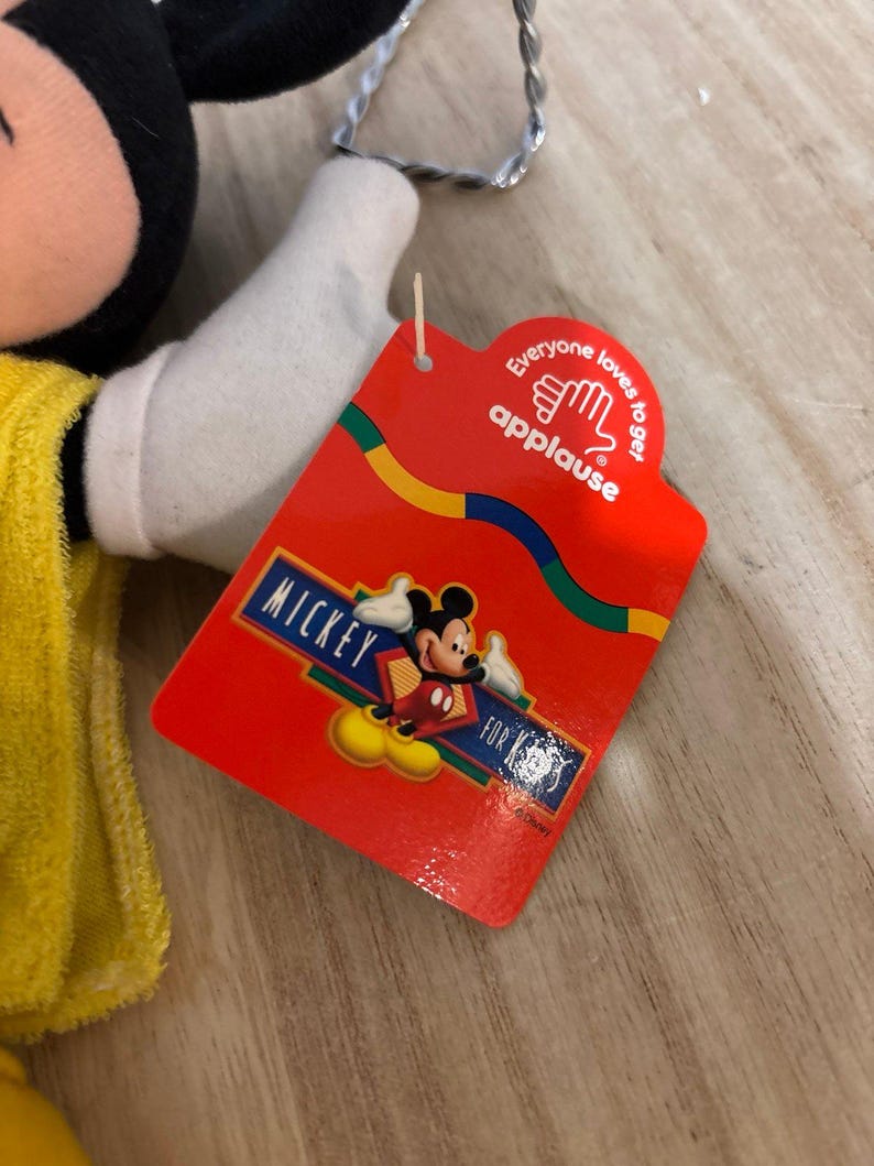 May include: A plush Mickey Mouse toy wearing a yellow garment, featuring a red cardboard tag attached by a plastic fastener. The tag displays the text "Everyone loves to get applause" and "MICKEY FOR KIDS" with a graphic of Mickey Mouse.