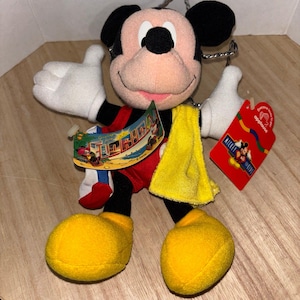 May include: A plush Mickey Mouse toy wearing red shorts and yellow shoes, featuring a yellow towel draped over one shoulder. The toy includes a "Greetings from Florida" tag and a red "Applause" brand tag with Mickey Mouse graphics attached.