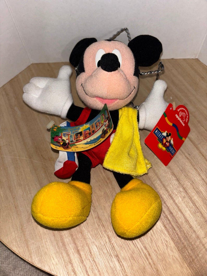 May include: A plush Mickey Mouse toy wearing red shorts and yellow shoes, featuring a yellow towel draped over one shoulder. The toy includes a "Greetings from Florida" tag and a red "Applause" brand tag with Mickey Mouse graphics attached.