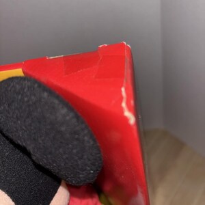 May include: A red box with a torn corner is partially visible. The box is next to a black plush ear and a red sleeve. The background is a light gray wall.