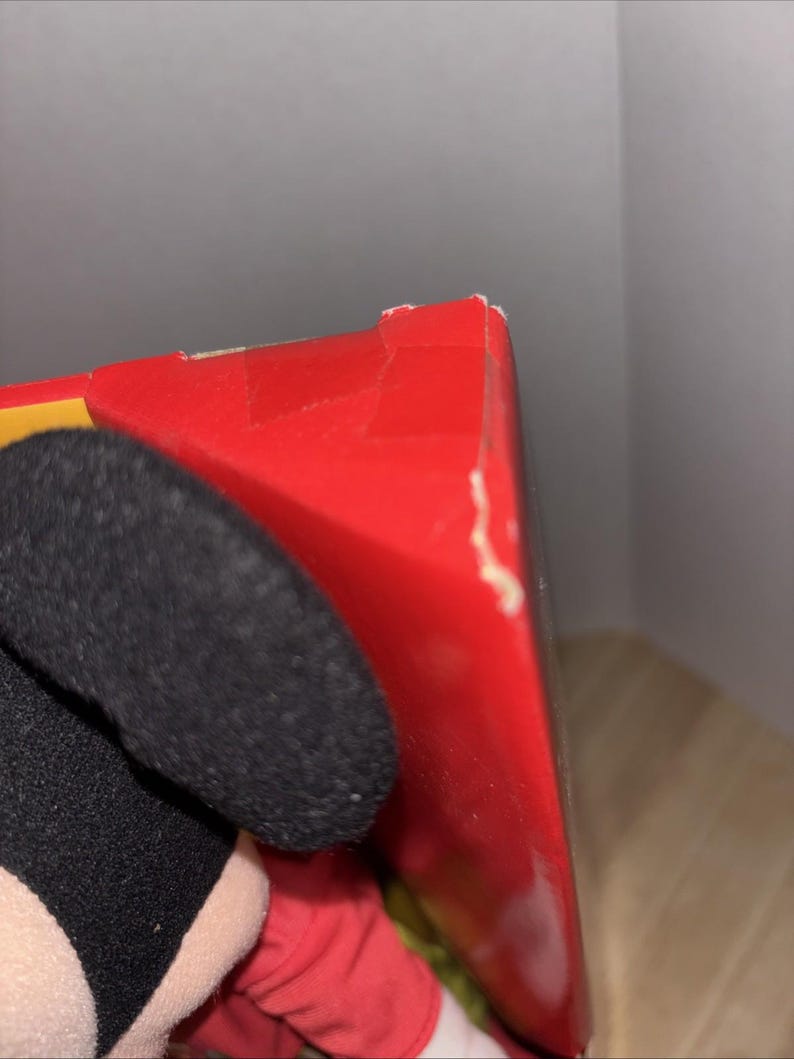 May include: A red box with a torn corner is partially visible. The box is next to a black plush ear and a red sleeve. The background is a light gray wall.