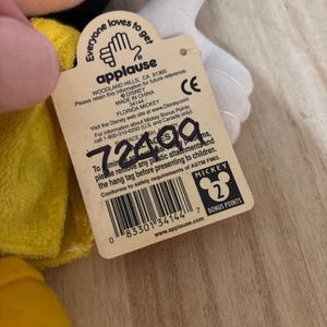May include: A beige cardboard hang tag attached to a plush toy, featuring the Applause logo, Disney branding, and the text Florida Mickey. The tag includes a barcode, safety information, and the handwritten number 72499 in black ink.