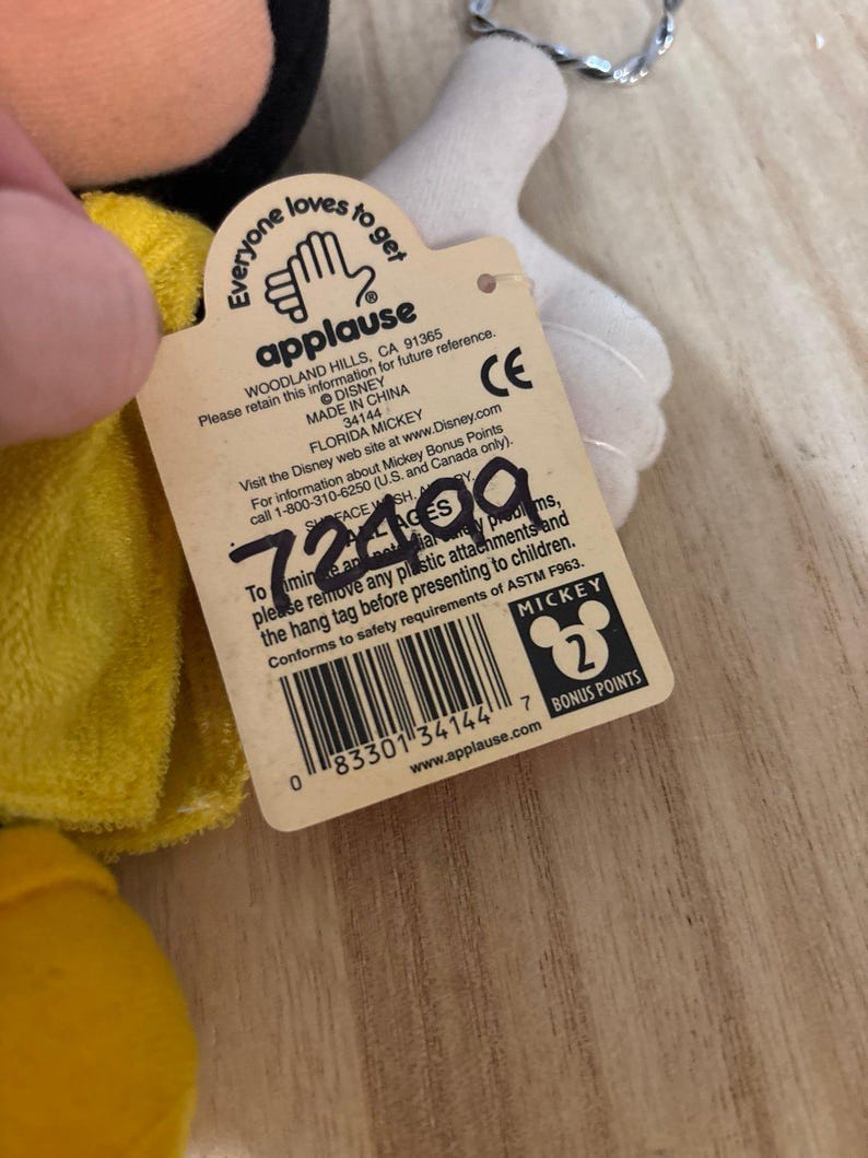 May include: A beige cardboard hang tag attached to a plush toy, featuring the Applause logo, Disney branding, and the text Florida Mickey. The tag includes a barcode, safety information, and the handwritten number 72499 in black ink.