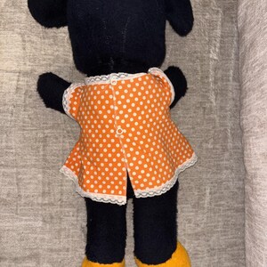 May include: A black plush doll with large ears, wearing an orange polka dot dress with white lace trim. The doll has yellow feet and black arms and legs. The back of the doll is facing the camera.