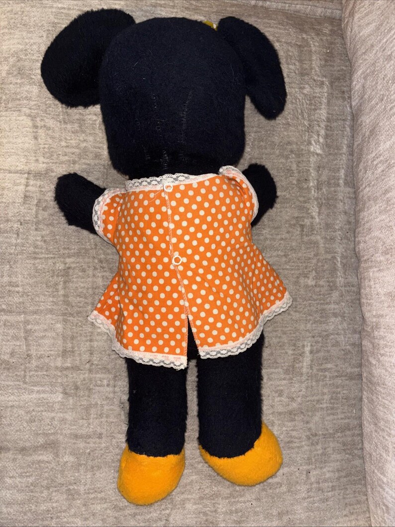 May include: A black plush doll with large ears, wearing an orange polka dot dress with white lace trim. The doll has yellow feet and black arms and legs. The back of the doll is facing the camera.
