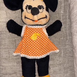 May include: A vintage Minnie Mouse plush doll. The doll has black fur, a cream-colored face, and a yellow bow. She wears an orange dress with white polka dots and a white lace trim. Her shoes are yellow.