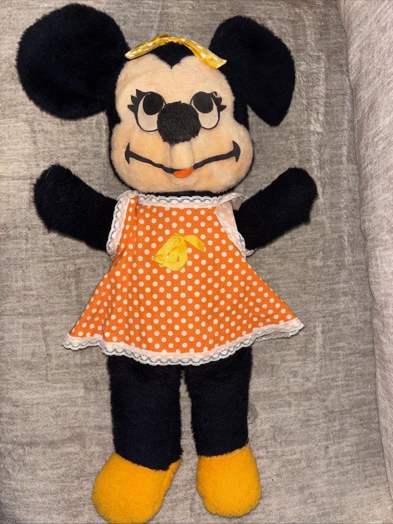 May include: A vintage Minnie Mouse plush doll. The doll has black fur, a cream-colored face, and a yellow bow. She wears an orange dress with white polka dots and a white lace trim. Her shoes are yellow.
