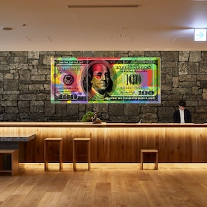 May include: A colorful, abstract design of a $100 US bill hangs on a stone wall above a wooden reception desk with two stools. The bill features a portrait of Benjamin Franklin.