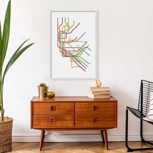 May include: A framed print of a colorful subway map, featuring lines in red, green, yellow, blue, and purple. The artwork is displayed on a white background and framed in white. The print is displayed above a wooden dresser.