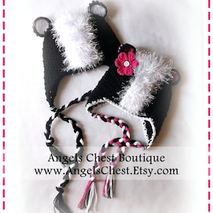 May include: Two black and white crocheted skunk hats with white fur trim and black and white braided ties. One hat has a pink flower with a button center.