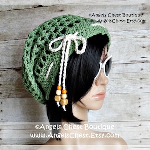 May include: Green crochet slouchy hat with a white crocheted bow and wooden bead accents. The hat is worn by a mannequin with black hair and white sunglasses. The text "Angels Chest Boutique www.AngelsChest.Etsy.com" is visible in the image.