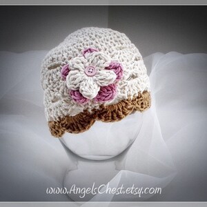 Może przedstawiać: Biały dziecięcy kapelusz na szydełku z brązowym brzegiem na szydełku i różową i białą kwiatem na szydełku z guzikiem w środku.