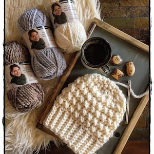 May include: Three skeins of yarn in different colors, a white crocheted hat, a cup of coffee, and wooden buttons on a wooden tray. The yarn labels read "Cozy Wool Laine Double Lana Accogliente".