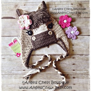 May include: Crocheted horse hat with brown and beige yarn, featuring a brown nose, black eyes, and a brown mane. The hat has a long brown and white braided cord. The hat is decorated with pink and purple flowers.