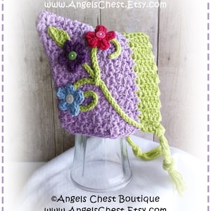 May include: A crocheted baby hat with a purple and green color scheme. The hat features a floral design with green leaves and colorful flowers. The hat has a long green tassel.