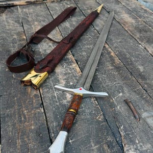Op de afbeelding: Een stalen zwaard met een tweesnijdend lemmet, een zilveren pareerstang met een kleine rode steen en een bruin gesneden houten handvat met gouden accenten. Het zwaard bevat een bijpassende bruine lederen schede met gouden metalen fittingen.