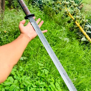 May include: A long, straight-edged sword with a dark, textured metal blade and a cylindrical handle featuring alternating dark brown and black bands. The sword has a simple, circular black guard separating the handle from the blade.