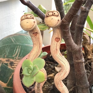 May include: Two whimsical ceramic plant stakes shaped like smiling worms with heart details. The stakes are tan with textured lines and are placed in a terracotta pot with succulents and other plants. The stakes add a playful touch to any garden.