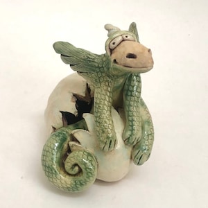 May include: A ceramic sculpture of a small green dragon emerging from a cracked white egg. The dragon has textured scales, large eyes, and a curled tail resting on the base. The piece features a glossy finish with green and white tones.