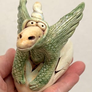May include: A ceramic sculpture of a small green dragon emerging from a cracked white egg. The dragon has textured scales, large eyes, and outstretched wings. The egg is smooth and off-white with jagged cracks where the creature appears.