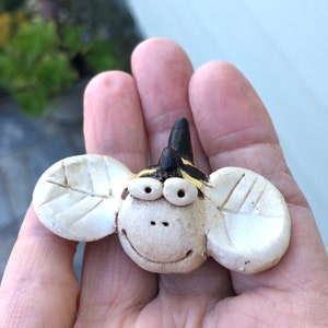 May include: A whimsical ceramic bee figurine with a smiling face, large eyes, and leaf-shaped wings. The bee has a black and yellow striped head and is held in a hand.