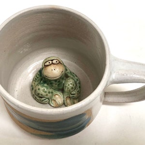 May include: A handmade ceramic mug with a light gray exterior and a whimsical green monkey figurine inside. The mug features a curved handle and a blue and tan wave design around the base. A unique and artistic piece for coffee or tea.