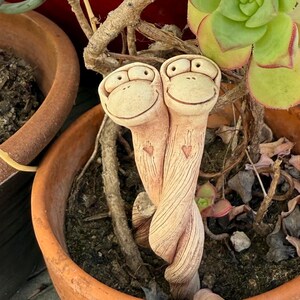 May include: A pair of whimsical ceramic monkey figurines with smiling faces, intertwined and planted in a terracotta pot. The monkeys are beige with etched details, nestled among succulents and dried branches. The pot is filled with soil and other plants.