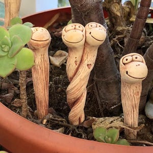 May include: A collection of whimsical, hand-crafted ceramic plant stakes. Each stake features a smiling monkey face and a textured, brown body. Two stakes are intertwined, with a heart carved into the design. The stakes are placed in a terracotta pot with green succulents.