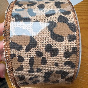 May include: A brown and black burlap ribbon with a leopard print design. The ribbon is wrapped around a clear plastic spool.
