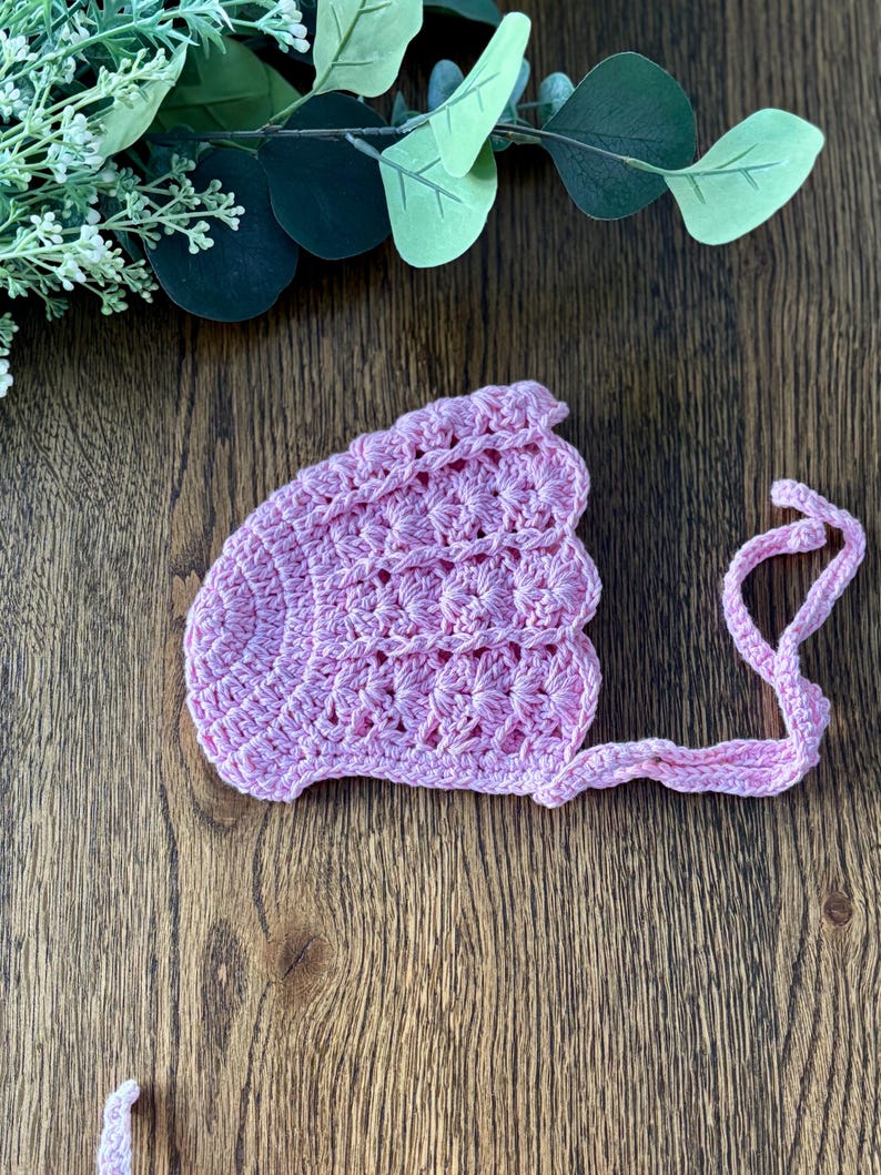 May include: A pink crocheted baby bonnet with a scalloped edge and a tie.