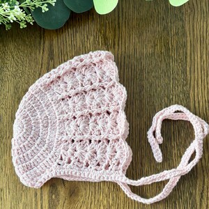 May include: A pink crocheted baby bonnet with a scalloped edge and ties. The bonnet is made with a delicate stitch pattern.
