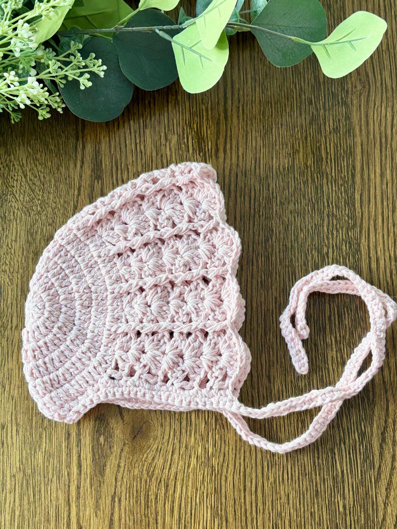 May include: A pink crocheted baby bonnet with a scalloped edge and ties. The bonnet is made with a delicate stitch pattern.