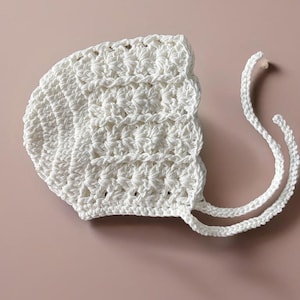 May include: White crocheted baby bonnet with two long ties. The bonnet has a scalloped edge and a textured stitch pattern. @ellenjoyce_crochet