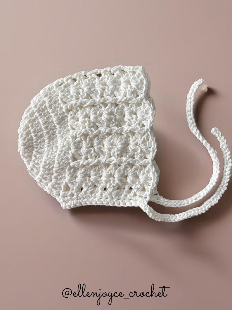 May include: White crocheted baby bonnet with two long ties. The bonnet has a scalloped edge and a textured stitch pattern. @ellenjoyce_crochet