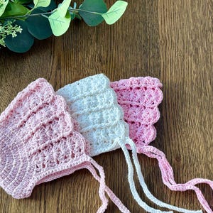 May include: Three crocheted baby bonnets in pink and white with ties. The bonnets are made with a delicate stitch pattern and have a scalloped edge.