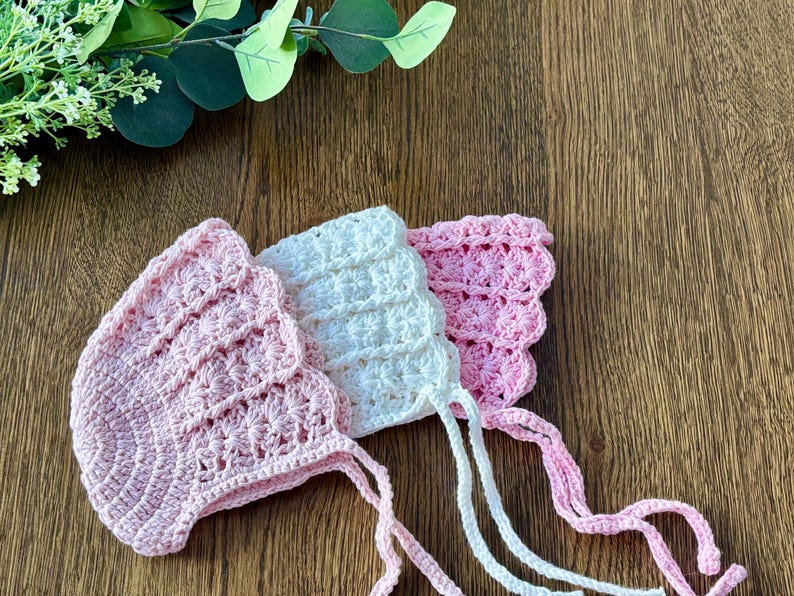 May include: Three crocheted baby bonnets in pink and white with ties. The bonnets are made with a delicate stitch pattern and have a scalloped edge.
