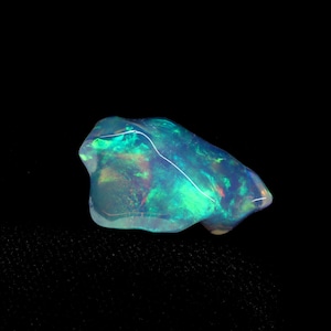 May include: A polished, irregular-shaped opal gemstone featuring a translucent body with vibrant flashes of neon green, electric blue, and subtle hints of red and orange light play throughout the stone against a dark background.