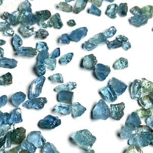 Puede incluir: Una colección de pequeños fragmentos de piedras preciosas en bruto, translúcidas, de color azul claro y verde pálido, esparcidos sobre una superficie blanca lisa. Cada pieza presenta bordes irregulares y una textura cristalina.