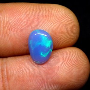 Könnte beinhalten: Ein oval geschliffener Opal-Edelstein. Der Opal zeigt eine lebendige blaue Grundfarbe mit wirbelnden Mustern aus Türkis und Grün. Der Edelstein wird zwischen zwei Fingern gehalten, um seine Größe und Farbvariationen zu zeigen.