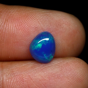 Peut inclure: Une pierre précieuse ovale polie, d'un bleu profond avec des reflets verts. La gemme est tenue entre deux doigts, mettant en valeur sa couleur vive et sa surface réfléchissante. Il s'agit probablement d'une opale.