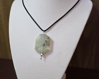 May include: A light green gemstone pendant necklace. The pendant is a faceted, irregular shape with dark inclusions, suspended from a black cord. The necklace is displayed on a white bust against a brown background.