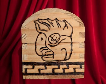 Könnte beinhalten: Eine helle Holzschachtel mit abgerundeter Oberseite, die auf der Vorderseite ein dunkles, eingebranntes Design eines stilisierten Gesichts aufweist. Unter dem Gesicht befindet sich ein horizontales Bordürenmuster, das ebenfalls in das Holz eingebrannt ist.