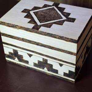 May include: A wooden box with a geometric design. The box is light-colored with dark brown accents. The lid features a central diamond shape surrounded by a stepped pattern. The sides have a repeating triangular design.