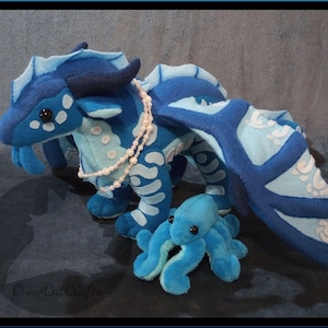May include: A plush blue dragon with white detailing and a pearl necklace, next to a small blue octopus plush. The image includes text that reads "SeaWing Plush Pattern Accessory Edition" and "Includes mini pearl tutorial and Octopus Plush Pattern."