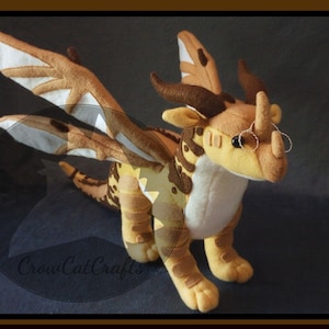 May include: A plush HiveWing dragon with tan, brown, and white wings and body. The dragon has small glasses and is part of a PDF sewing pattern. The image includes text that says "CrowCatCrafts" and "HiveWing Plush Pattern".