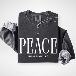 May include: Dark gray long-sleeved shirt with the word "PEACE" in large white letters, a cross, and the text "PHILIPPIANS 4:7". A silver necklace with a pendant is draped over the shirt. The sleeves are cuffed.