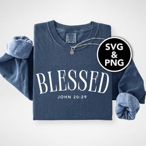 May include: A navy blue sweatshirt with the word "BLESSED" in large white letters and the text "JOHN 20:29" below. The sleeves are cuffed, and a silver necklace with a pendant is visible. A black and white SVG & PNG graphic is in the image.