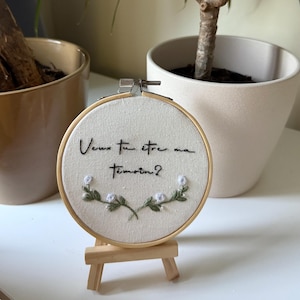 Könnte beinhalten: Ein runder Stickrahmen mit dem französischen Text "Veux tu être ma témoin?" in schwarzem Faden. Die Stickerei zeigt ein Blumendesign mit weißen Blüten und grünen Blättern. Der Rahmen wird auf einer kleinen Holzstaffelei präsentiert.