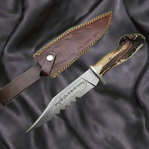 May include: A hunting knife with a serrated blade and a handle made of antler. The knife is accompanied by a brown leather sheath. The blade has etched text. The knife is displayed on a dark, textured surface.