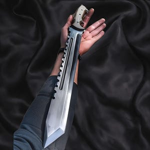 May include: A large, polished silver knife with a serrated edge and a pointed tip. The knife has a light-coloured handle and a wide blade. The blade is held in a person's hand against a black background.
